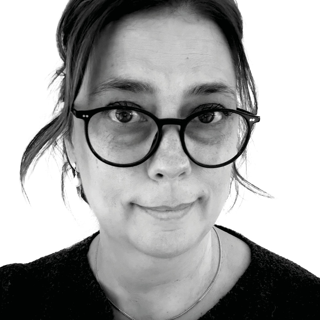 Frau mit Brille und dunklem Haar, die einen Pullover und eine Halskette trägt, vor einem weißen Hintergrund.