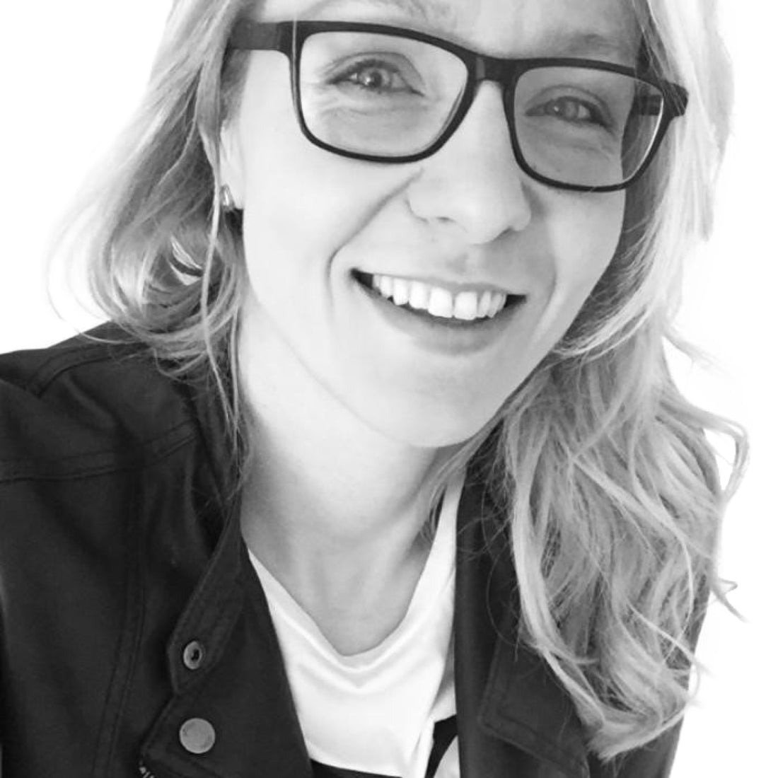 Lächelnde Frau mit Brille und gewelltem Haar, die eine Jacke trägt und in die Kamera schaut.