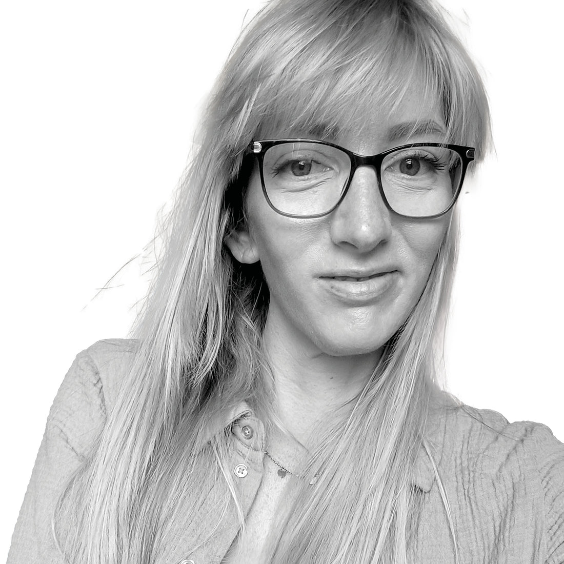 Eine Frau mit langem blondem Haar und Brille lächelt leicht, sie trägt ein Hemd mit Knopfleiste auf einem Schwarz-Weiß-Foto.