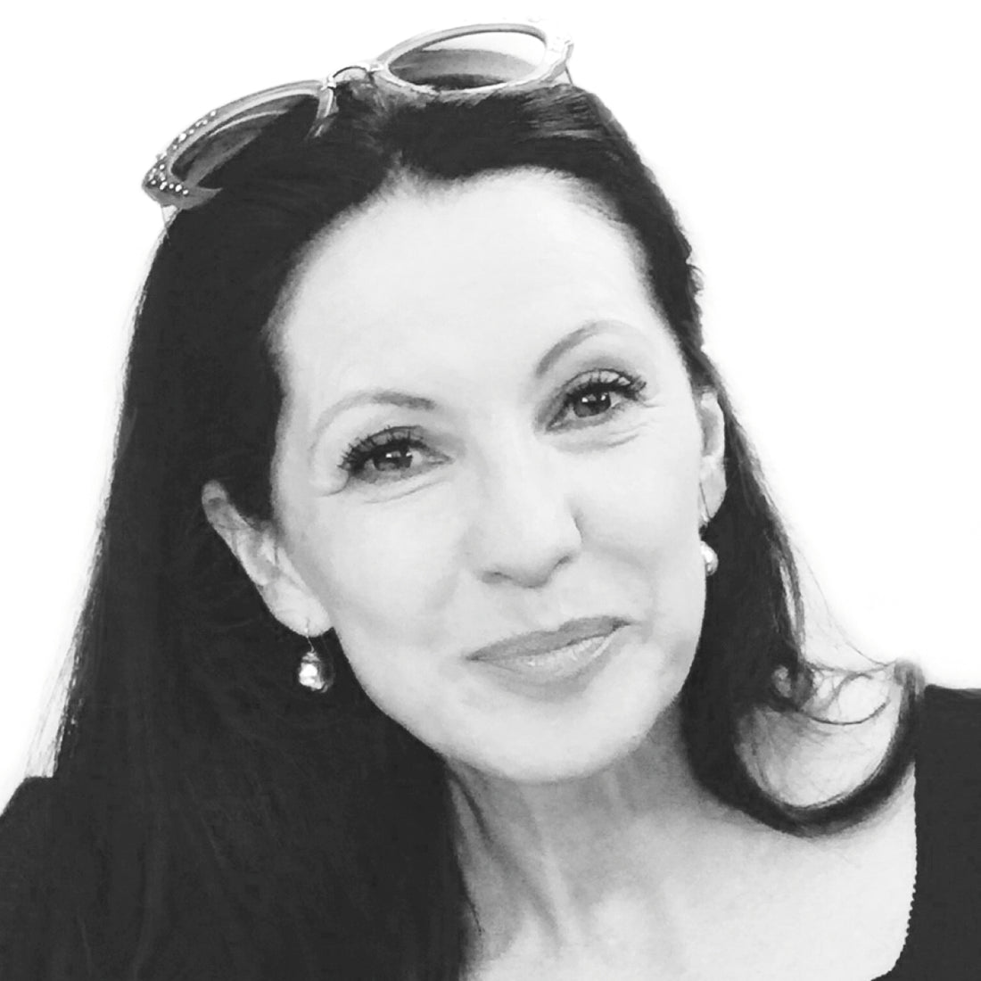 Lächelnde Frau mit langen dunklen Haaren, die eine Sonnenbrille auf dem Kopf und Perlenohrringe trägt, in Schwarz und Weiß.