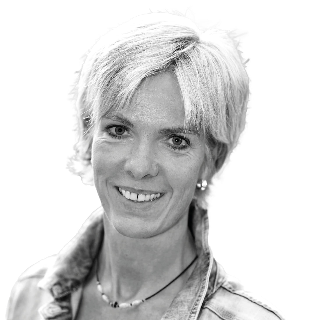 Frau mit kurzen blonden Haaren, lächelnd, trägt eine Jeansjacke und eine Halskette, auf weißem Hintergrund.