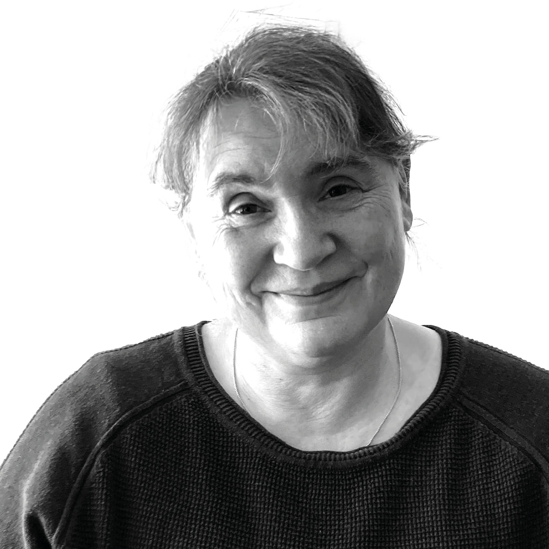 Lächelnde ältere Frau mit grauem Haar, die einen dunklen Pullover trägt, fotografiert vor einem weißen Hintergrund.