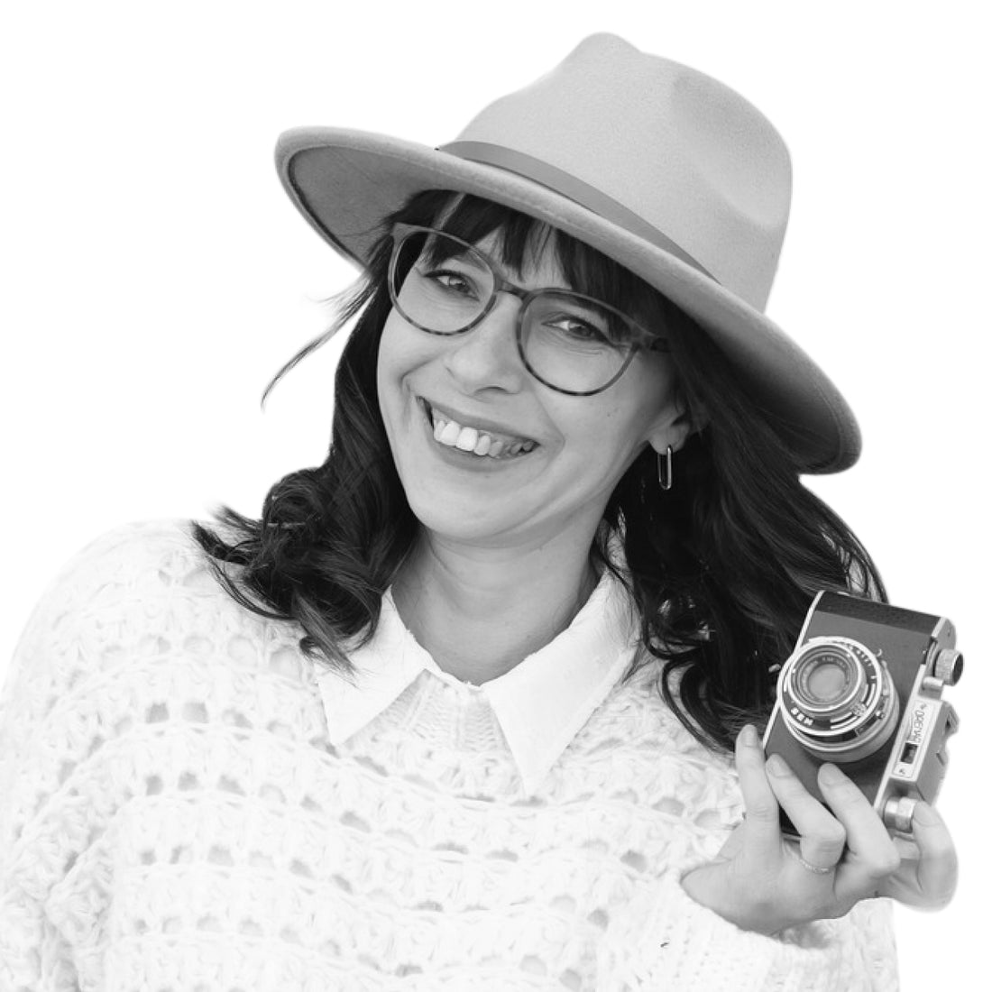 Lächelnde Frau mit Brille und Hut, die eine Vintage-Kamera hält und einen weißen Pullover trägt, auf weißem Hintergrund.