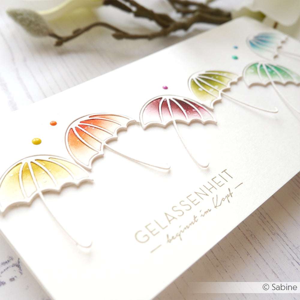 Eine Karte mit bunten, geprägten Regenschirmen aus der Alexandra Renke Motivstanze Regenschirm und dem in Gold gedruckten Wort "Gelassenheit".