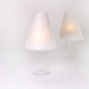 Zwei Weingläser mit dekorativen Tischlampen von Alexandra Renke, die jeweils mit transparenten Papierlampenschirmen versehen sind, leuchten sanft vor einem schlichten weißen Hintergrund.