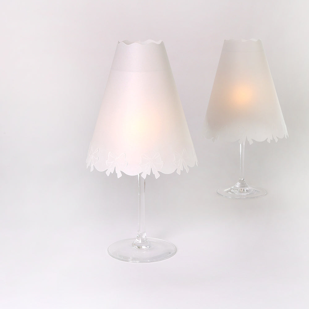 Zwei Weingläser mit dekorativen Tischlampen von Alexandra Renke, die jeweils mit transparenten Papierlampenschirmen versehen sind, leuchten sanft vor einem schlichten weißen Hintergrund.