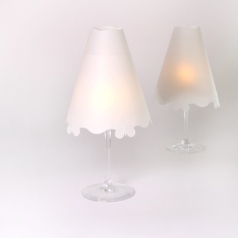 Zwei Weingläser mit sanft leuchtenden weißen Alexandra Renke Transparentpapier-Lampenschirmen auf schlichtem weißen Hintergrund - perfekt als stilvolle Tischdekoration.