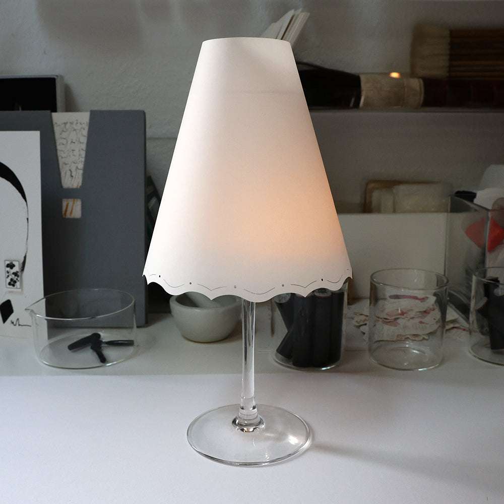 Ein Alexandra Renke Weinglas mit einem Lampenschirm aus Papier auf der Oberseite schafft einen eleganten, kleinen Tischleuchten-Look und dient als stilvolle Tischdeko auf einem weißen Schreibtisch.