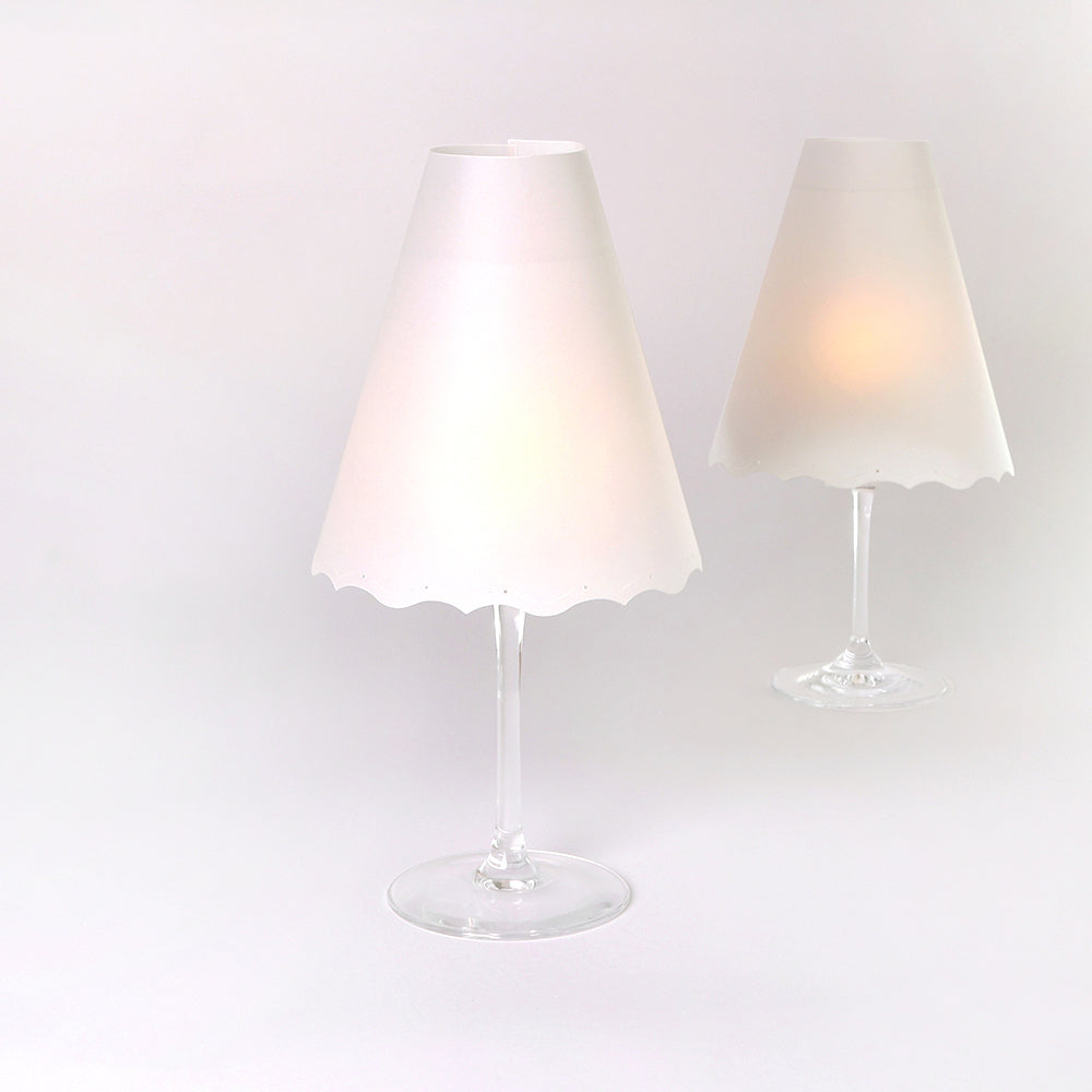 Zwei Weingläser mit weißen Lampenschirmdeckeln aus Lampenschirme Transparentpapier von Alexandra Renke, die an kleine Tischleuchten Deko erinnern, stehen auf einem schlichten Hintergrund.