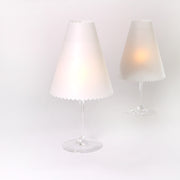 Zwei Weingläser mit weißen Lampenschirmen aus Transparentpapier von Alexandra Renke, die eleganten Tischlampen ähneln und sanft leuchten, bilden eine charmante Tischdeko auf weißem Hintergrund.