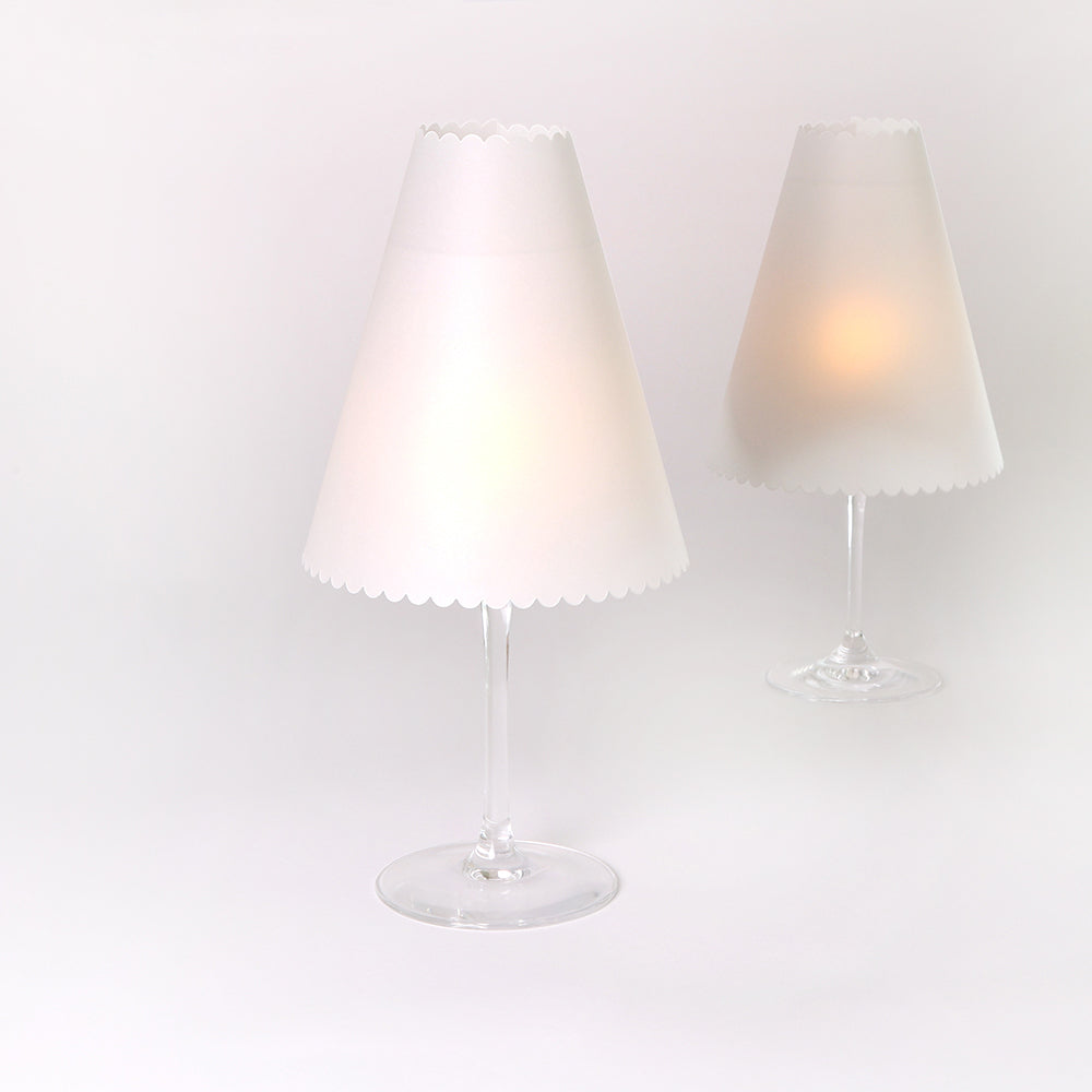 Zwei Weingläser mit weißen Lampenschirmen aus Transparentpapier von Alexandra Renke, die eleganten Tischlampen ähneln und sanft leuchten, bilden eine charmante Tischdeko auf weißem Hintergrund.