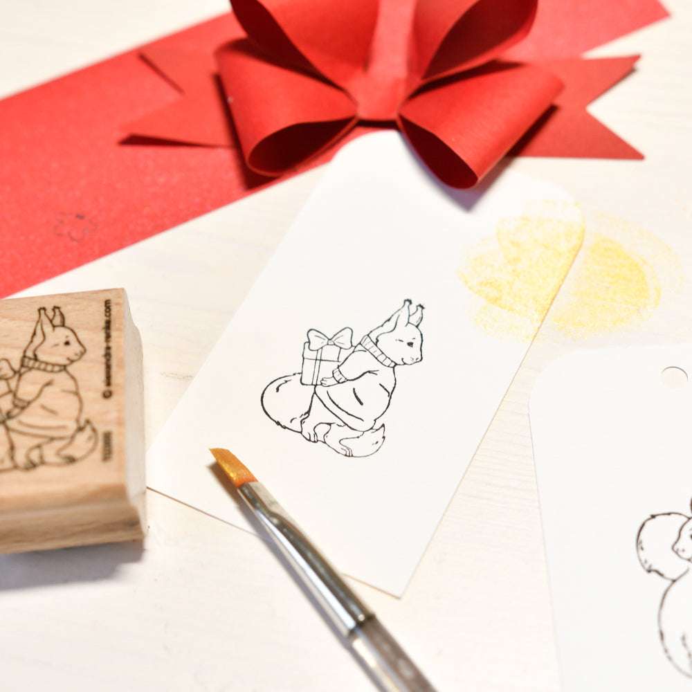 Ein hochwertiger Holzstempel von Alexandra Renke zeigt ein Eichhörnchen mit Geschenk, Pinsel, roter Schleife und Goldglitter auf weißem Hintergrund.