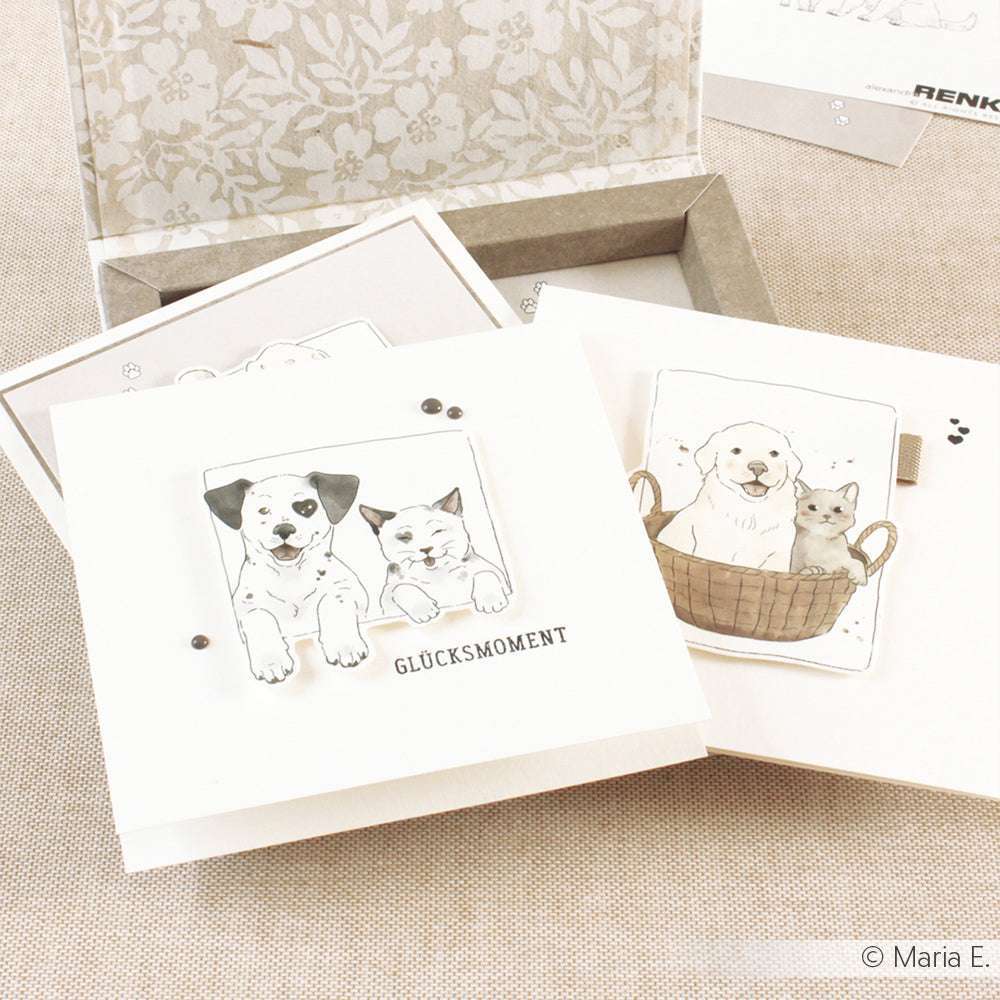 Drei Alexandra Renke-Grußkarten mit Hunde- und Katzen-Illustrationen, darunter eine mit "GLÜCKSMOMENT", auf einer beigen Oberfläche in warmem Grau - ideal für die kreative Kartengestaltung.