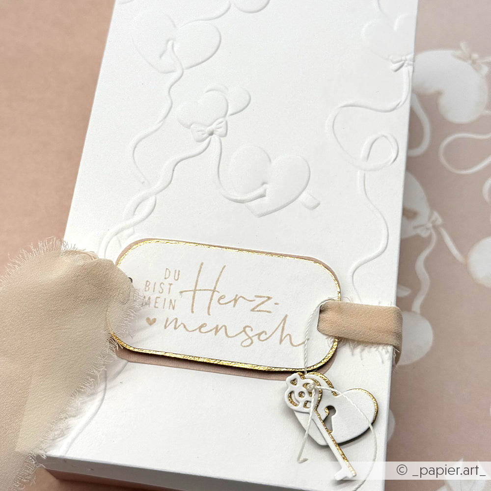 Weiße Geschenkbox von Alexandra Renke, hergestellt mit Designpapier und Prägefolder. Mit geprägten Verbandelten Herzen, einer Schleife, einem "Du bist mein Herzmensch"-Anhänger und einem kleinen Schlüsselanhänger.