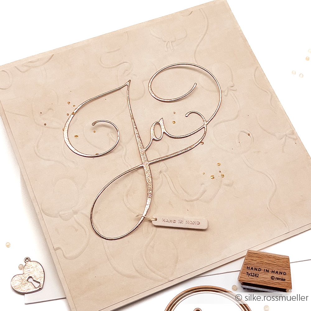 Quadratische beige Karte von Alexandra Renke mit elegantem Ja-Schriftzug, einem kleinen Hand in Hand-Anhänger und zarten Schmuckakzenten. Mit Papierprägetechniken für zusätzliche Textur und Charme.