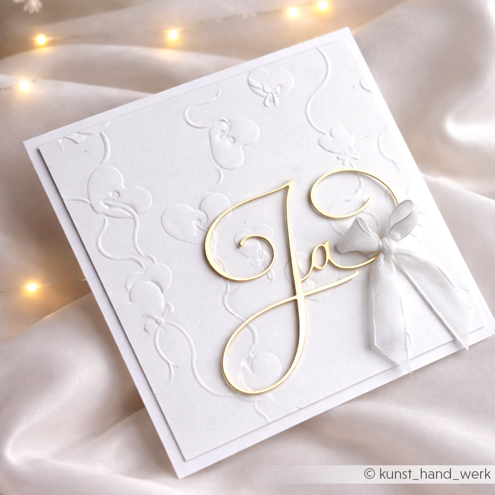 Weiße Prägekarte von Alexandra Renke mit Prägen Papier und goldenem "Ja"-Schriftzug, verziert mit einer weißen Bandschleife auf seidigem Stoff und sanften Lichtern.