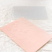 Eine rosa geprägte Karte mit Blumenmuster, die mit dem Alexandra Renke Embossing Folder hergestellt wurde, liegt neben einer klaren Prägeplatte auf zerknittertem weißen Papier.