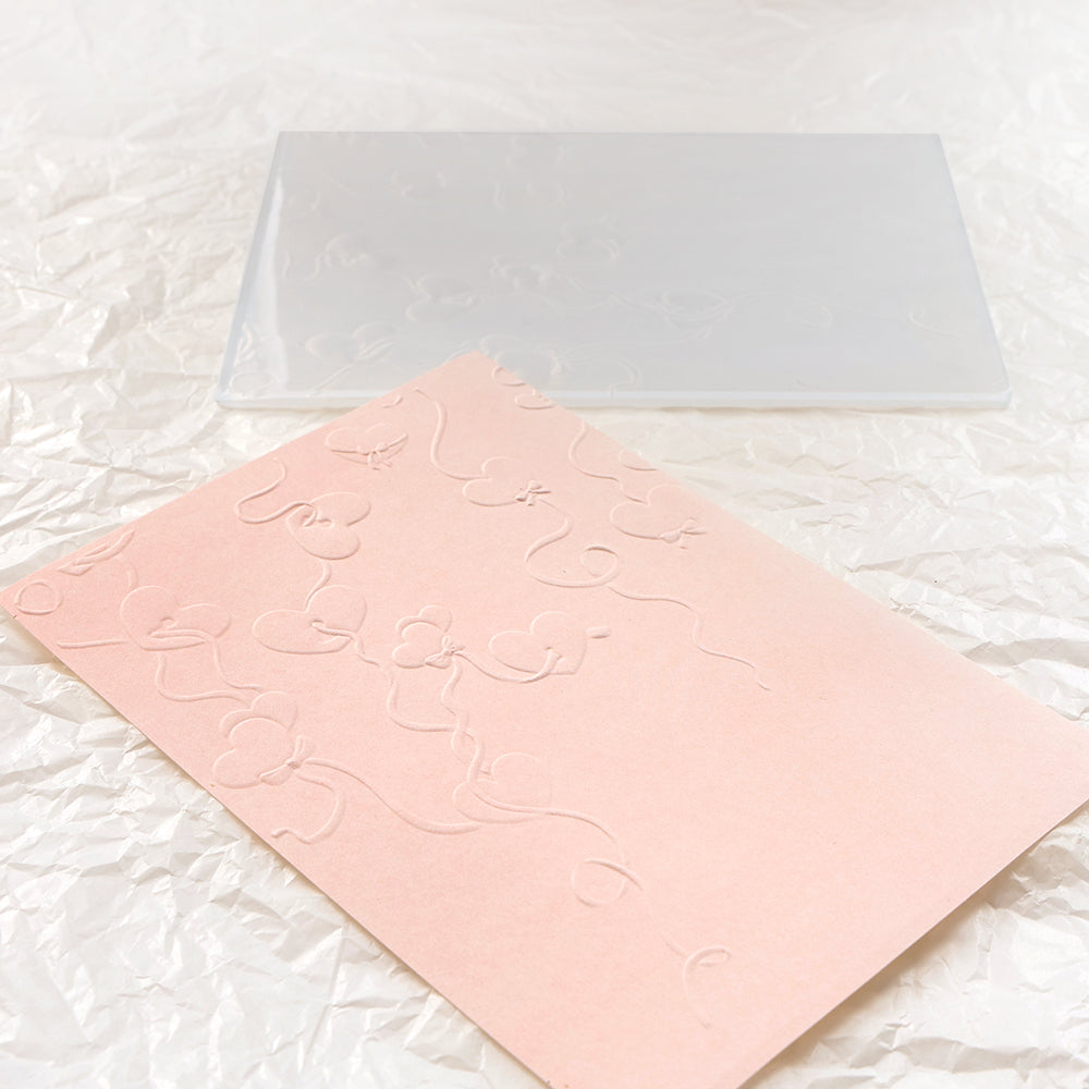 Eine rosa geprägte Karte mit Blumenmuster, die mit dem Alexandra Renke Embossing Folder hergestellt wurde, liegt neben einer klaren Prägeplatte auf zerknittertem weißen Papier.