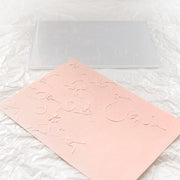 Eine rosa geprägte Karte, die mit dem Alexandra Renke Embossing Folder hergestellt wurde, und ein durchscheinender Umschlag liegen auf geknitterten weißen Alexandra Renke Designpapieren.