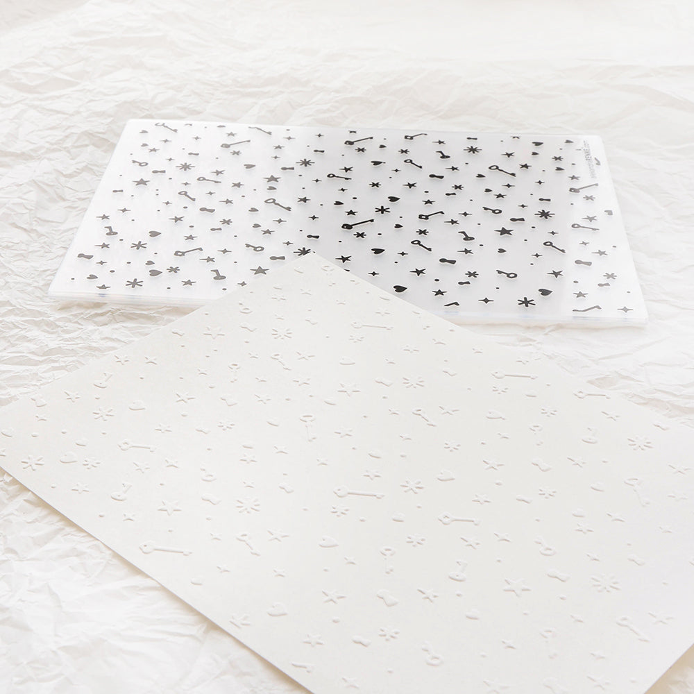 Ein weißes Blatt, das mit einem Alexandra Renke Cut & Emboss Folder mit Symbolen geprägt wurde, und ein klares Blatt mit passenden schwarzen Symbolen liegen auf einer strukturierten Oberfläche, die an Zauberhafte Schlüsselmagie erinnert.
