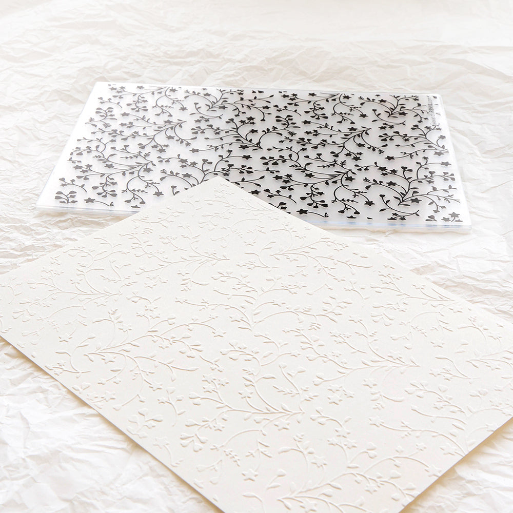 Alexandra Renke Cut & Emboss Folder aus durchsichtigem Kunststoff mit einem floralen Design, das neben geprägtem cremefarbenem Papier auf einem zerknitterten weißen Hintergrund gezeigt wird.