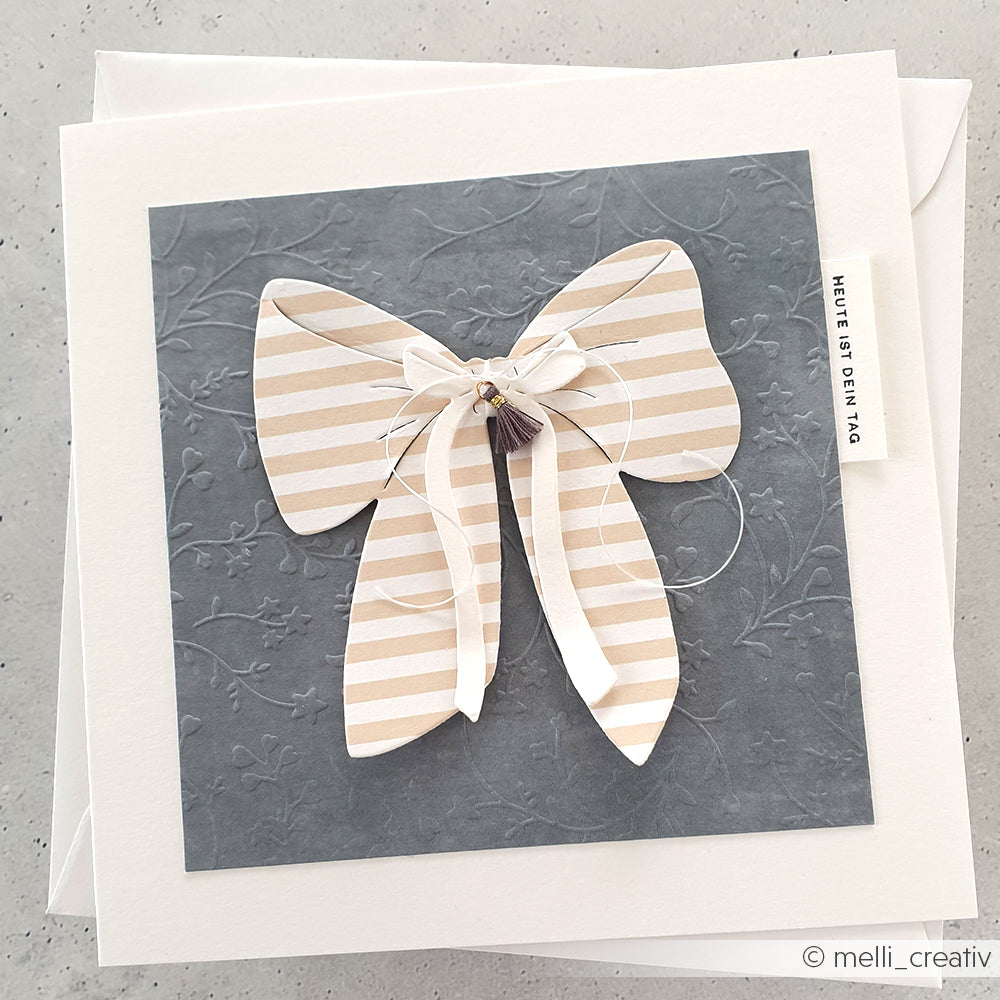 Die handgefertigte Karte von Alexandra Renke zeigt eine große gestreifte Schleife auf geprägtem grauen Papier, die mit dem Prägefolder Sternenranken hergestellt wurde, und ist mit einem kleinen "HEUTE IST DEIN TAG"-Anhänger versehen.