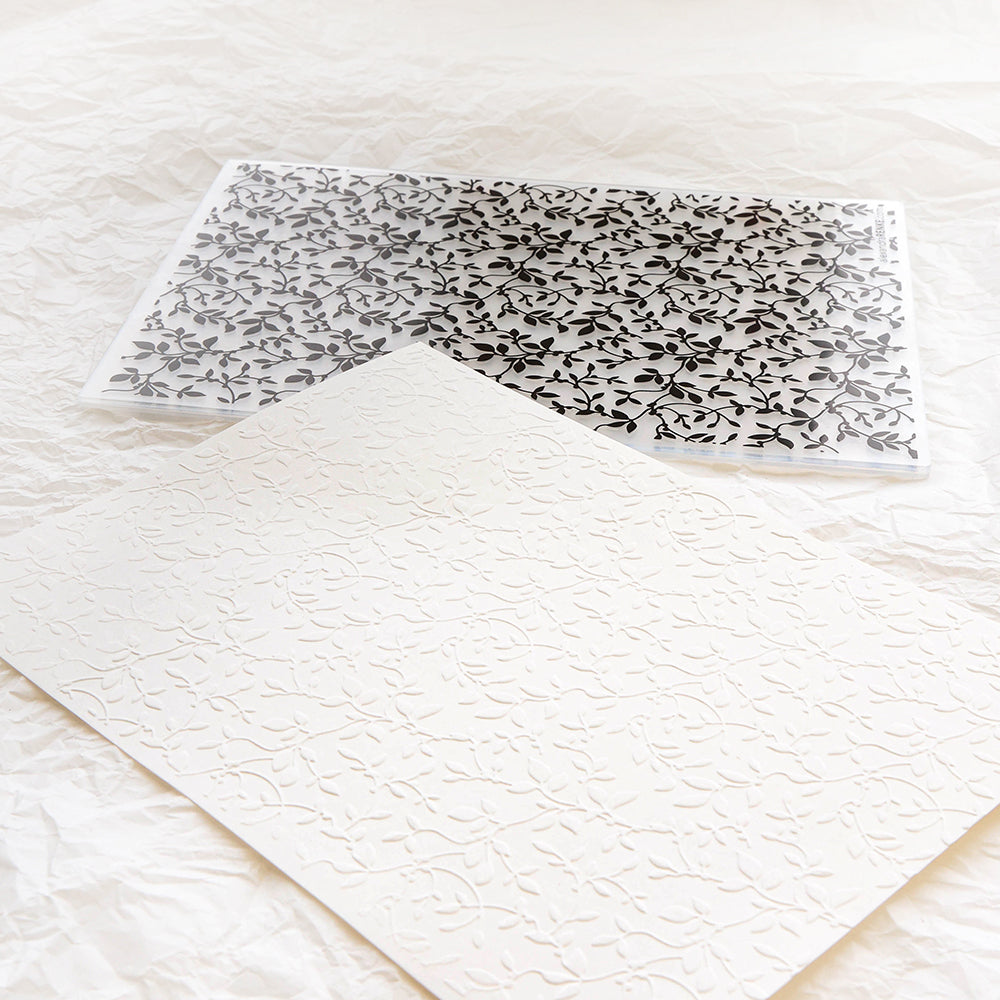 Ein weißes Blatt mit einem erhabenen Blattmuster, das mit dem Alexandra Renke Prägefolder Beerenzweige hergestellt wurde, liegt neben einer transparenten Schablone mit demselben eleganten Muster.