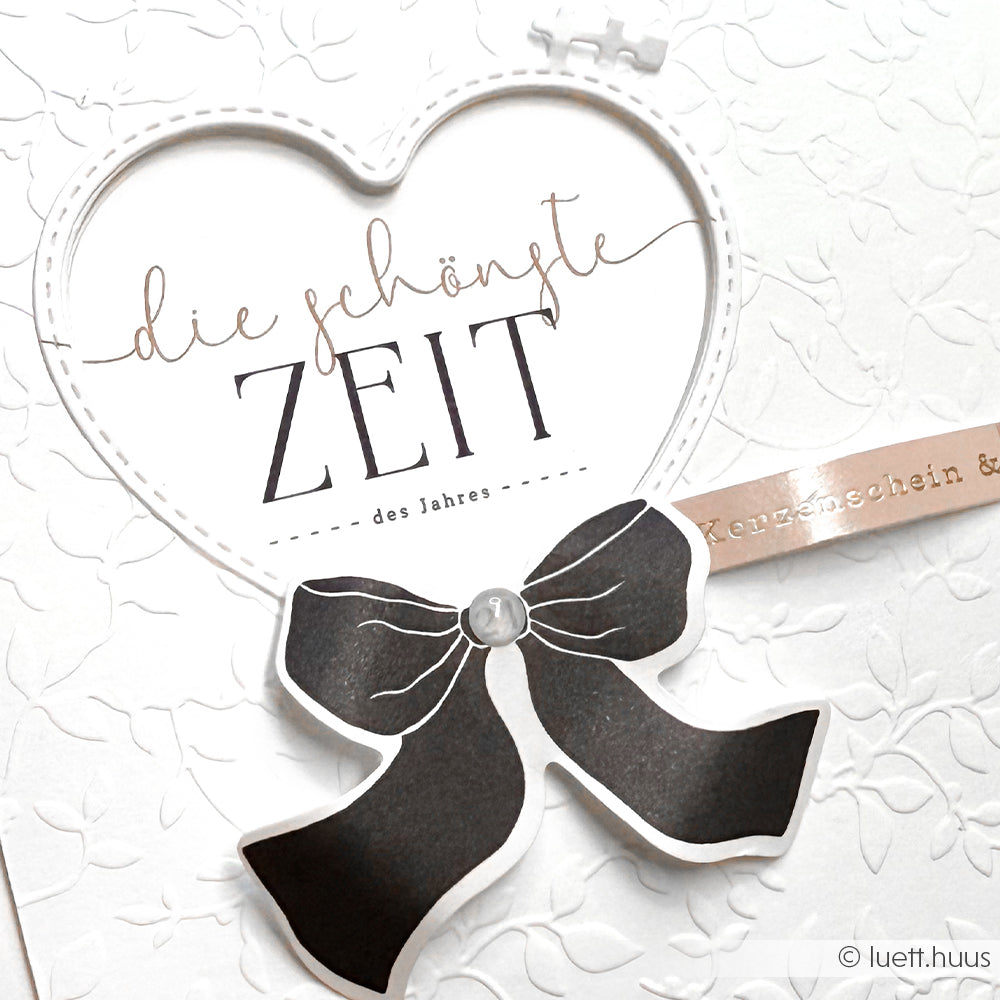 Weiße Karte mit einem herzförmigen Fenster, das mit dem Schneide- und Prägefolder von Alexandra Renke hergestellt wurde, mit dem Text "die schönste ZEIT" und einer schwarzen Schleifengrafik am unteren Rand.