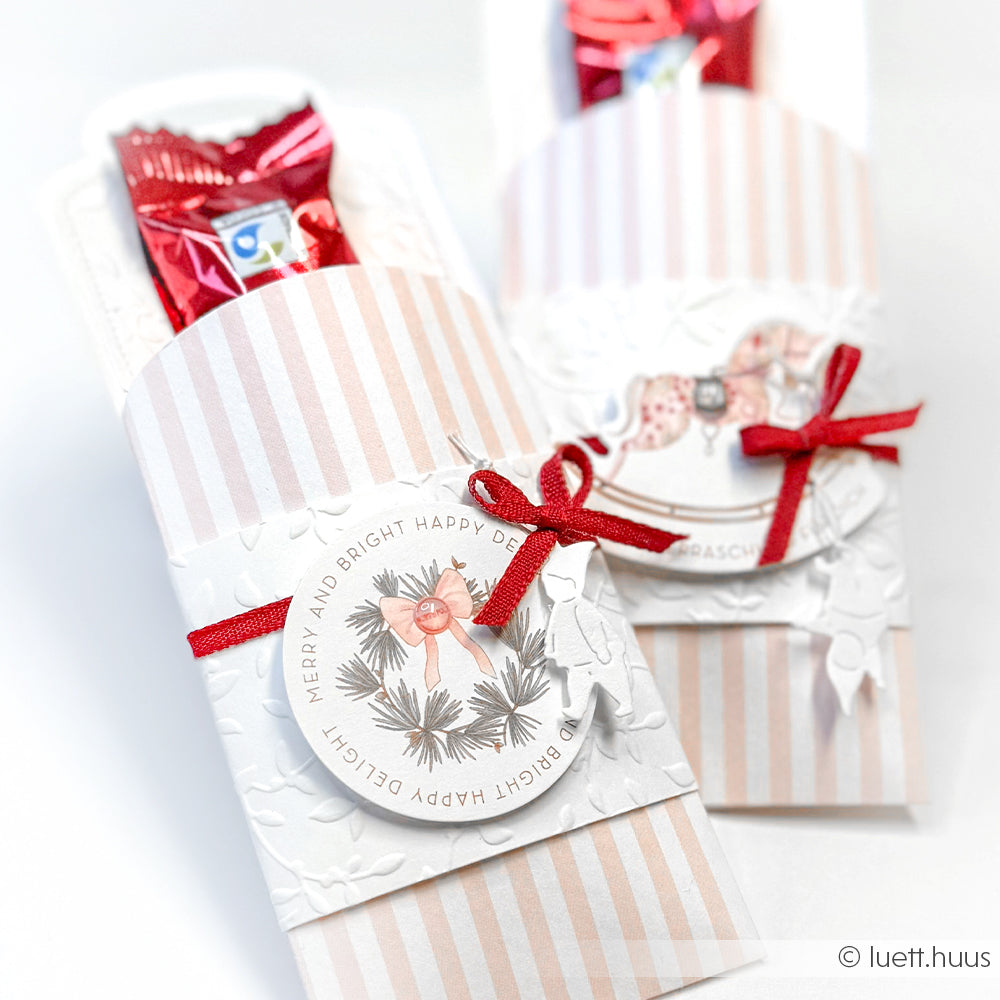 Zwei festliche Bonbonverpackungen mit roten Bändern und weihnachtlichen Anhängern von Alexandra Renke sind auf einer weißen Fläche arrangiert, die mit einem Prägefolder kunstvoll gestaltet ist.