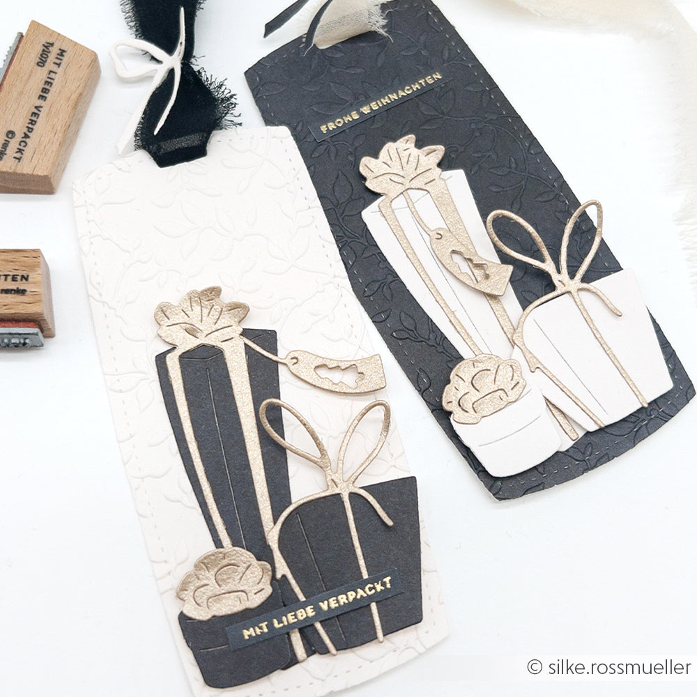 Zwei handgefertigte Geschenkanhänger von Alexandra Renke mit geprägten Geschenken - einer schwarz, einer weiß - mit goldenen Details und Bändern.