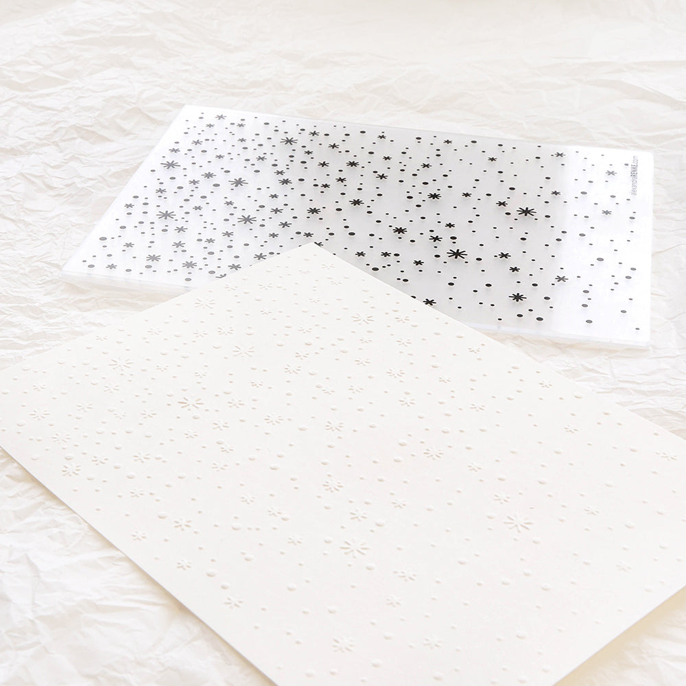 Ein weißes Blatt, das mit einem Schneide- und Prägefolder von Alexandra Renke mit Sternenmustern geprägt wurde, liegt neben einer transparenten Schablone mit denselben Sternenmustern.