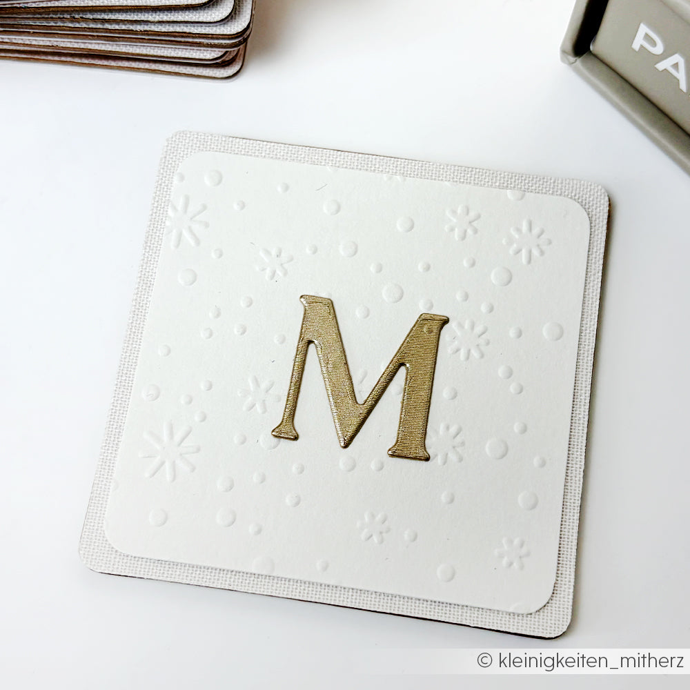 Eine weiße Karte mit dem goldenen Buchstaben M sowie geprägten Punkten und Schneeflocken, hergestellt mit dem Prägefolder Zarter Flockentanz von Alexandra Renke.