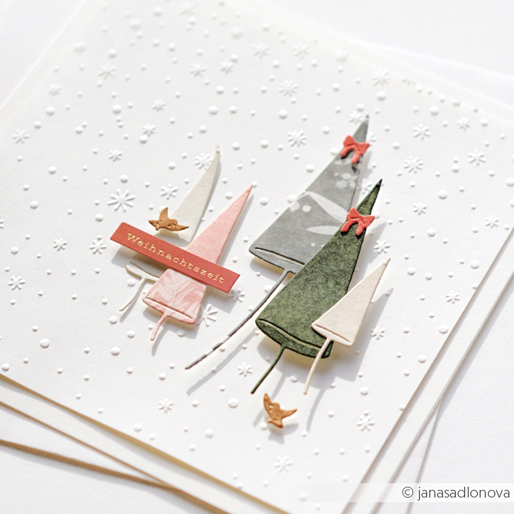 Minimalistische Weihnachtskarte mit geschichteten Papierbäumen, roten Schleifen, kleinen Vögeln und geprägten Schneeflocken, die mit dem Prägefolder Zarter Flockentanz von Alexandra Renke hergestellt wurden.