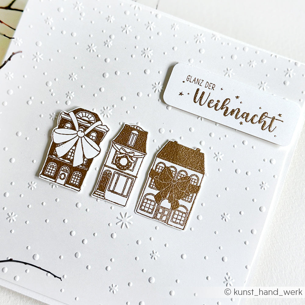 Weiße Weihnachtskarte von Alexandra Renke mit drei goldgeprägten Häusern und dem Text "Glanz der Weihnacht" auf verschneitem Hintergrund, erstellt mit dem Cut & Emboss Folder - perfekt, um den zarten Flockentanz der Saison zu feiern.