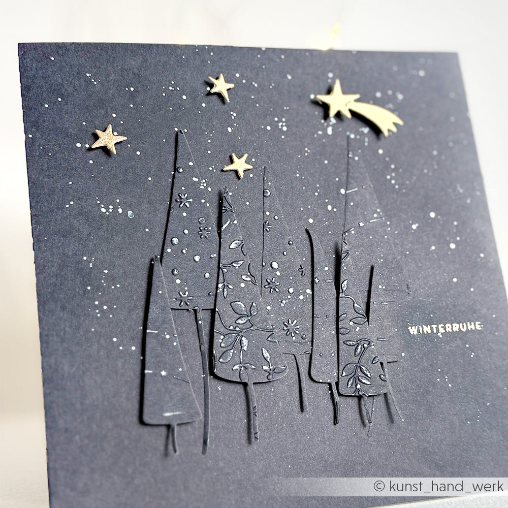 Handgemachte Karte von Alexandra Renke mit ausgeschnittenen Bäumen, die mit dem Cut & Emboss Folder hergestellt wurden, goldenen Sternen, einer Sternschnuppe und einem WINTERRUHE-Spruch auf dunklem Hintergrund.