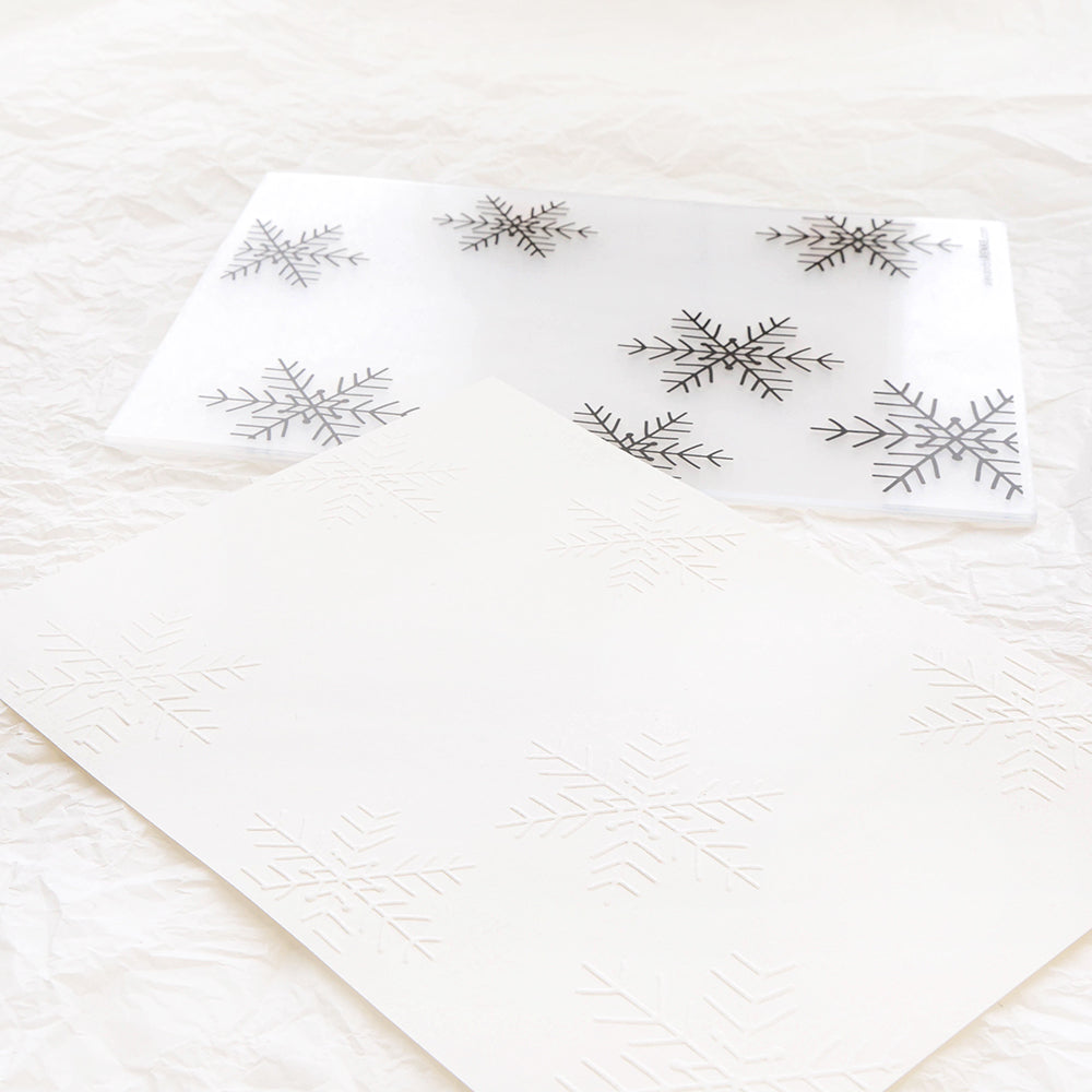 Zwei durchscheinende Blätter mit Großen Schneeflocken-Motiven von Alexandra Renke, die mit einem Prägefolder hergestellt wurden, liegen auf zerknittertem weißem Papier - ein Blatt zeigt schwarze Konturen.