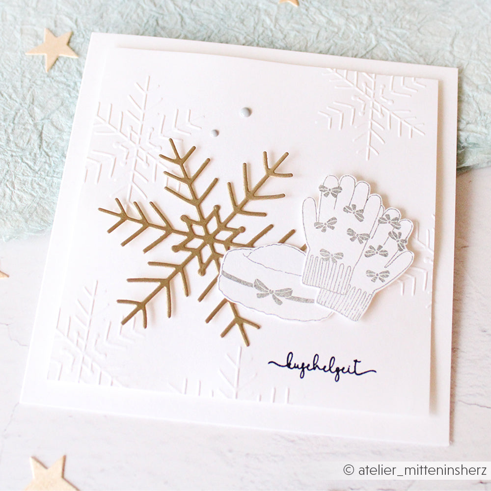 Quadratische weiße Karte von Alexandra Renke mit geprägten großen Schneeflocken, einer goldenen Schneeflocke, Fäustlingen und Ohrenschützern. Die Karte wurde mit einem Embossing Folder erstellt und enthält den Schriftzug "Herzlichkeit" für einen warmen Wintergruß.