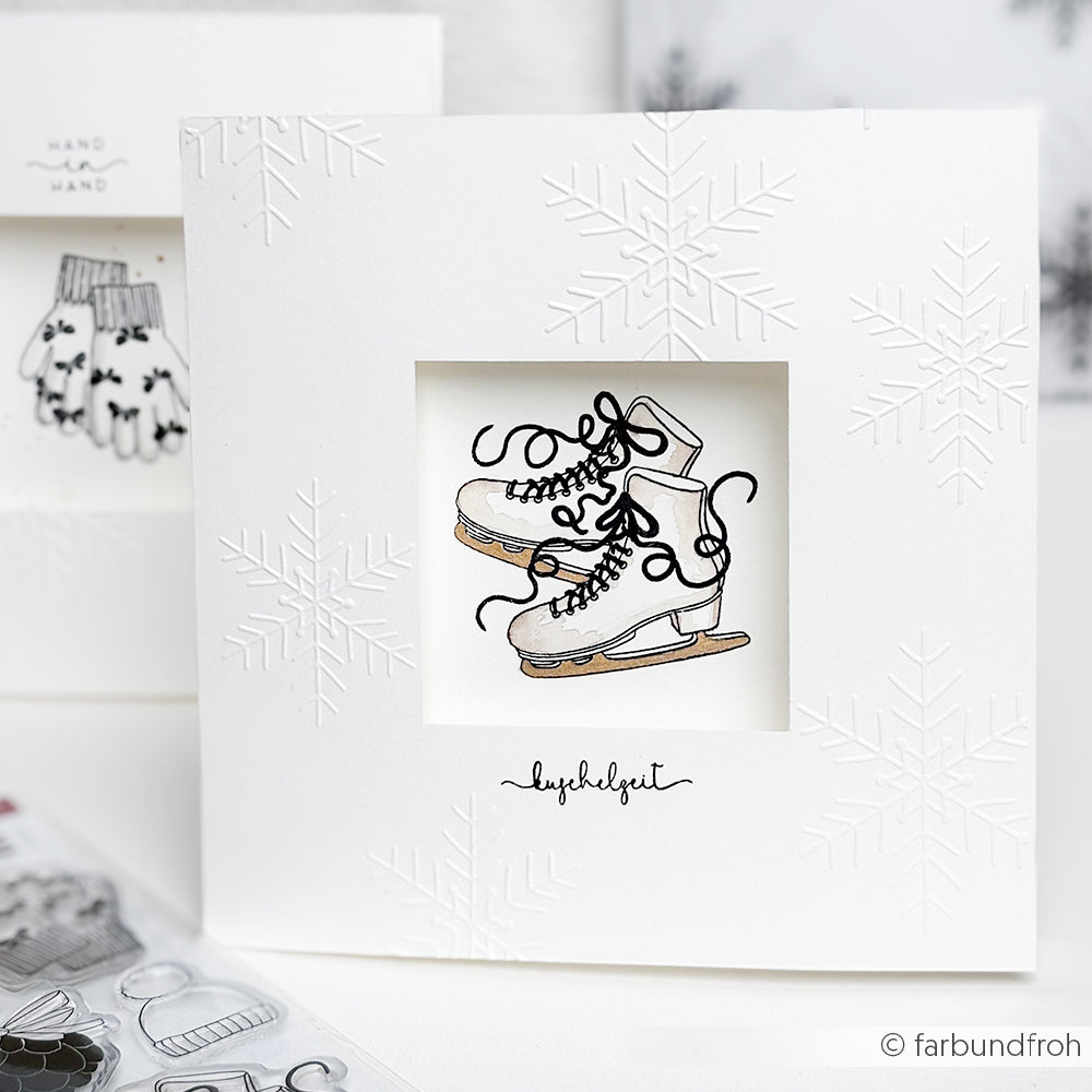 Weiße Karte mit geprägten Schneeflocken mit dem Schneide- und Prägefolder von Alexandra Renke, mit einer Zeichnung von Schlittschuhen, deren Spitzen durch ein quadratisches Fenster sichtbar sind.