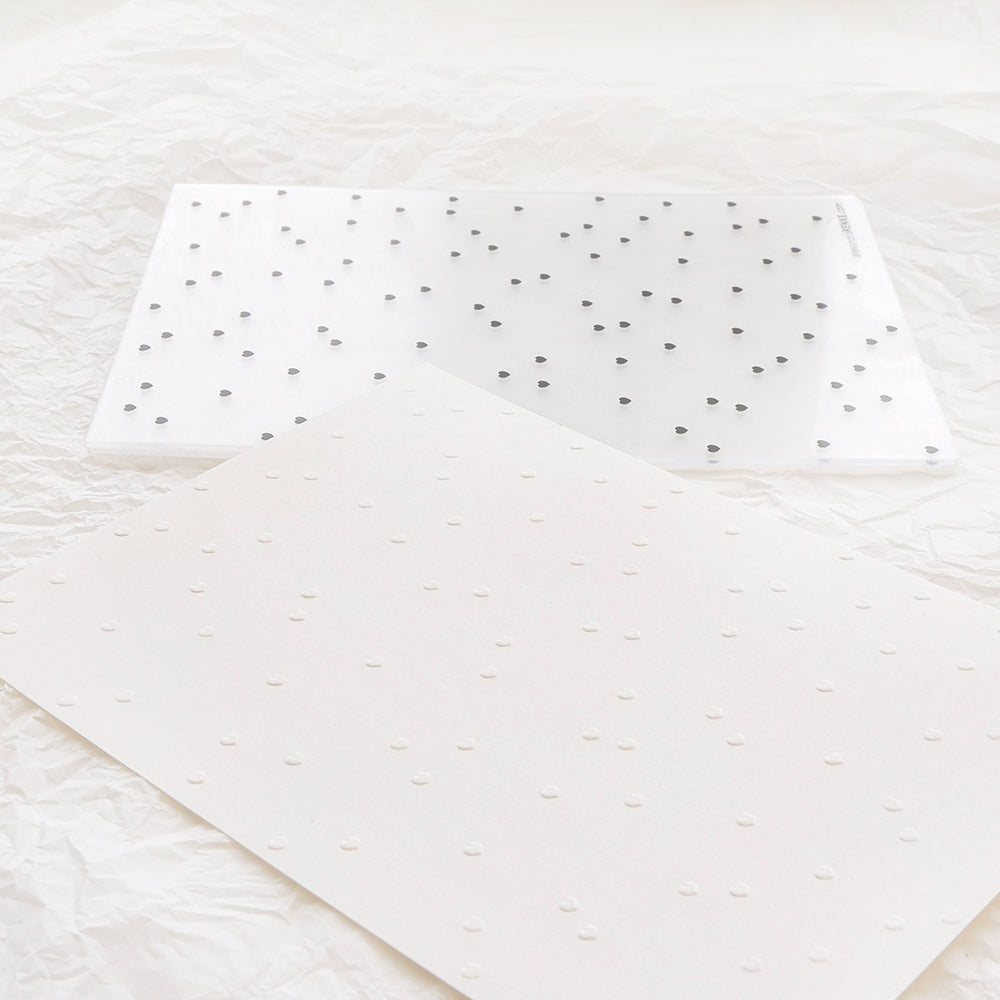 Ein weißer Prägefolder mit kleinen Punkten und passendem geprägtem weißem Papier, genannt Herzchenregen von Alexandra Renke, ausgestellt auf zerknittertem weißem Stoff.