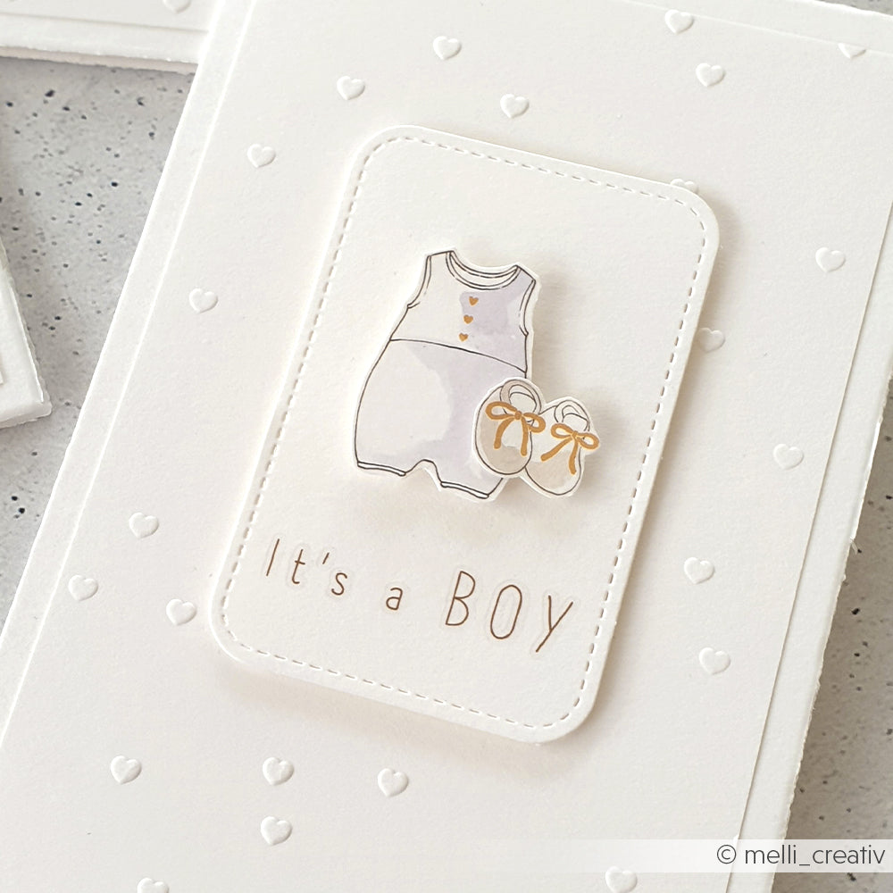 Weiße Karte mit einer Illustration eines Babystramplers und Schuhen, die mit einem Prägefolder von Alexandra Renke bearbeitet wurde, und "It's a BOY" in brauner Schrift.