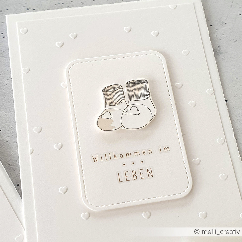 Weiße Karte von Alexandra Renke mit kleinen Herzen, Babysocken-Illustration und dem Text "Willkommen im Leben". Mit einem bezaubernden Herzchenregen-Motiv, das für zusätzliche Textur geprägt wurde.