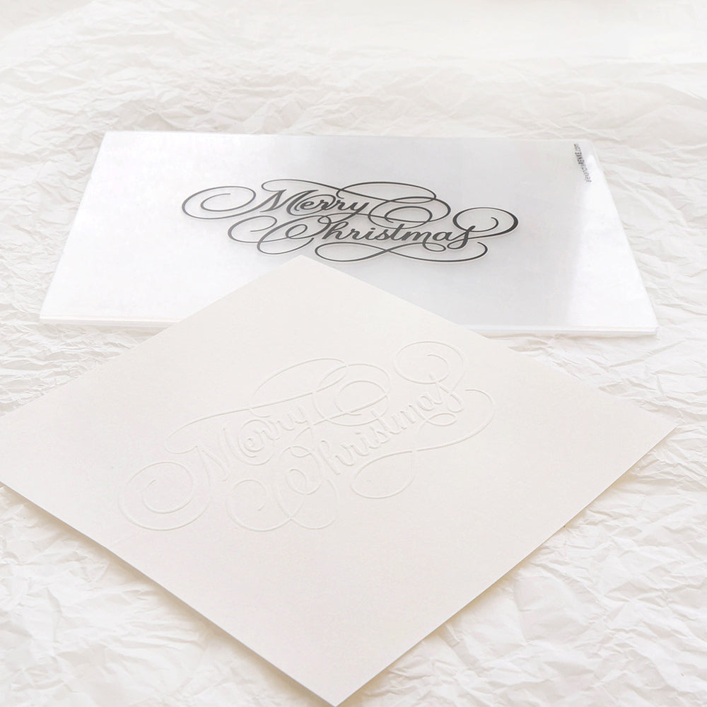 Weiße Karte von Alexandra Renke mit der Prägung "Frohe Weihnachten", die neben einem Embossing-Folder auf einem Hintergrund aus zerknittertem weißem Papier zu sehen ist.