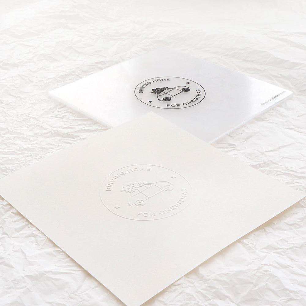 Zwei quadratische Drucke von Alexandra Renke - ein weißer mit geprägtem Text aus einem Cut & Emboss Folder und ein klarer mit schwarzer Tinte - liegen auf zerknittertem weißem Papier und erinnern an das gemütliche Gefühl, zu Weihnachten nach Hause zu fahren.
