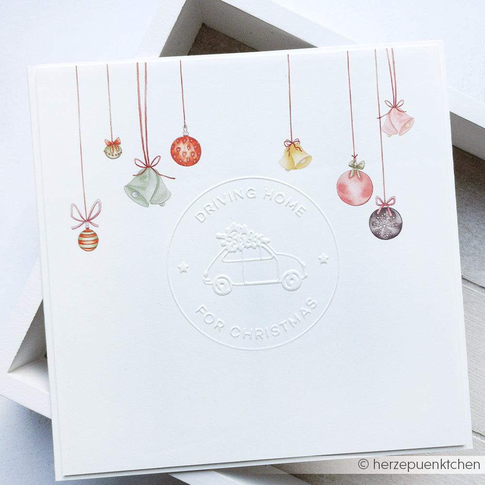 Diese weiße Karte von Alexandra Renke zeigt hängende Ornamente und Glocken, ein mit dem Cut & Emboss Folder geprägtes Auto mit einem Baum und den Schriftzug "Driving Home For Christmas".
