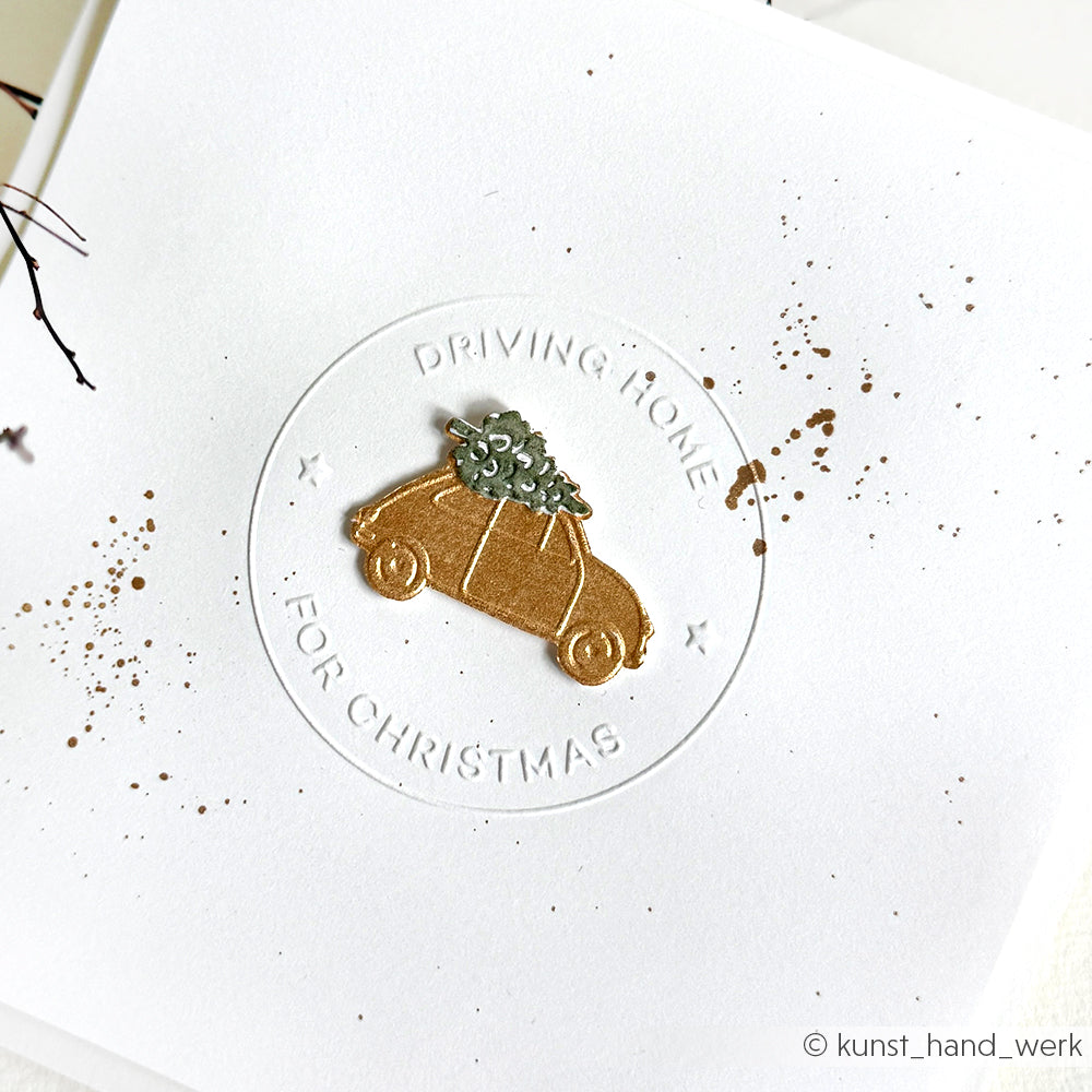 Ein goldenes Auto mit einem Weihnachtsbaum auf der Spitze wurde mit dem Schneide- und Prägefolder von Alexandra Renke auf eine weiße Karte geprägt und mit dem Text Driving Home for Christmas" versehen.