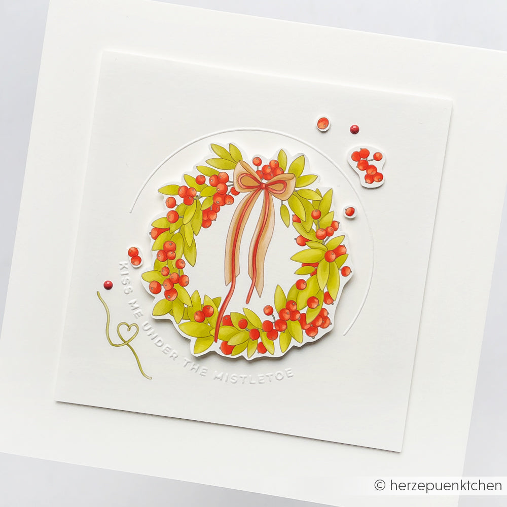 Eine festliche Karte von Alexandra Renke mit einem Kranz aus grünen Blättern, roten Beeren und einer Schleife, die mit dem Cut & Emboss Folder erstellt wurde, und dem Titel "Kiss Me Under the Mistletoe".