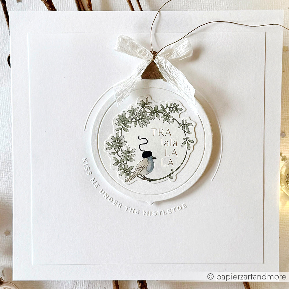 Weiße Karte mit einem kreisförmigen Anhänger mit Grünzeug, einem Vogel, "TRAlalaLALA" und einer Schleife; hergestellt mit dem Alexandra Renke Stanzsystem und mit geprägtem Text darunter.