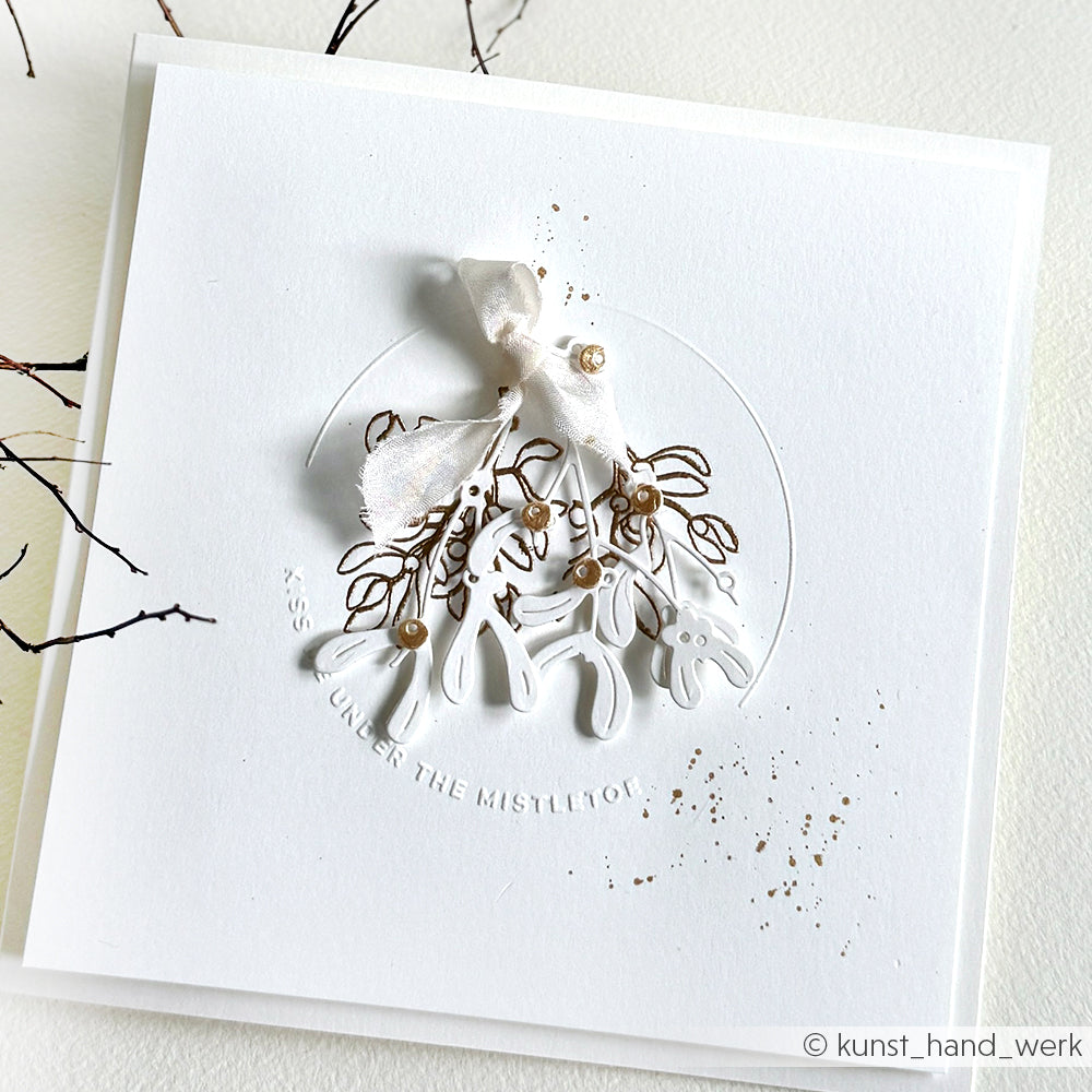 Minimalistische weiße Karte von Alexandra Renke, die mit einem Prägefolder bearbeitet wurde. Mit geprägtem Kreis, Mistelzweig, Schleife und goldenen Akzenten.