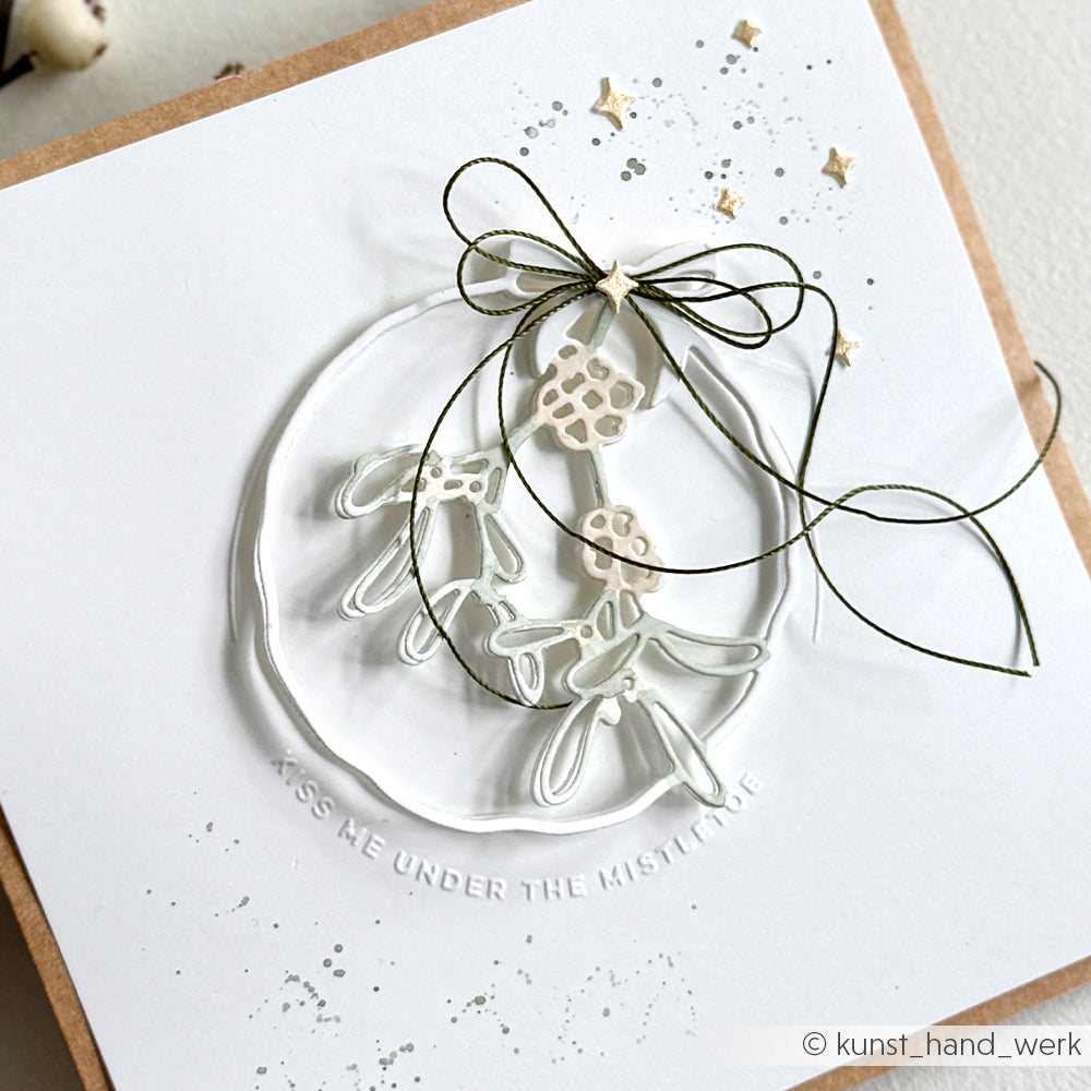 Handgefertigte Karte von Alexandra Renke mit einem Mistelzweig-Motiv, grüner Schleife und der Prägung "KISS ME UNDER MISTLETOE" unten, hergestellt mit einem Cut & Emboss Folder für zusätzliche Struktur und Details.
