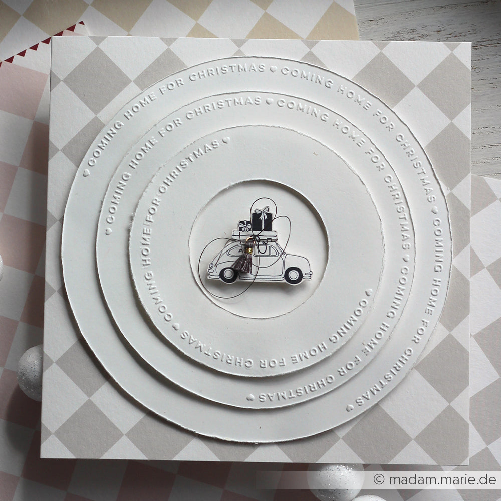 Weiße Karte von Alexandra Renke, gestaltet mit einem Prägefolder mit geprägten konzentrischen Kreisen, einer kleinen Autoillustration in der Mitte und Weihnachtsgeschenken auf dem Dach.